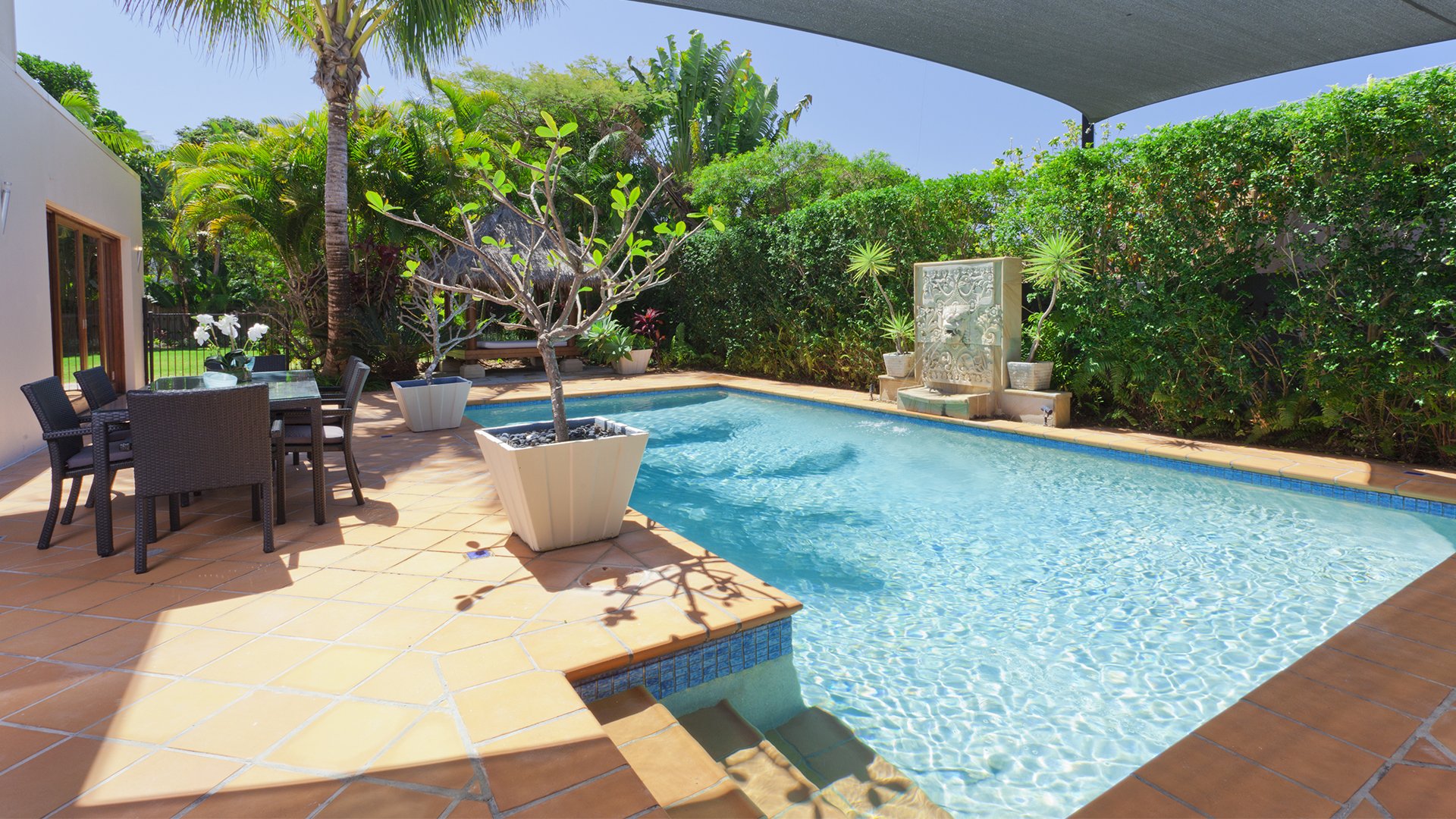 Outdoor swimming pool beside a tiled patio with dining furniture, tropical plants, and a stone fountain.