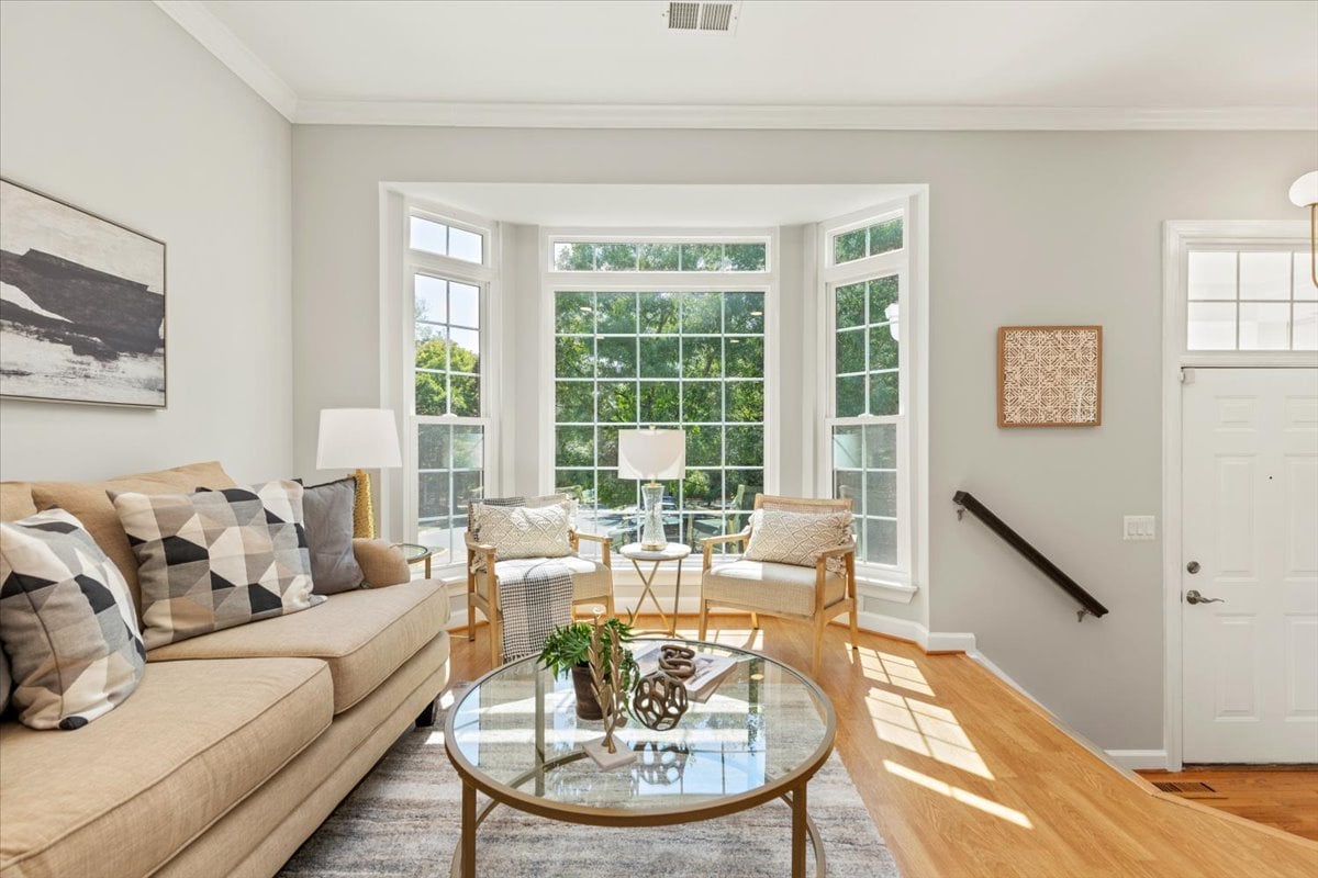 Living room with a beige sectional and seating arranged beside a large, sunlit bay window.