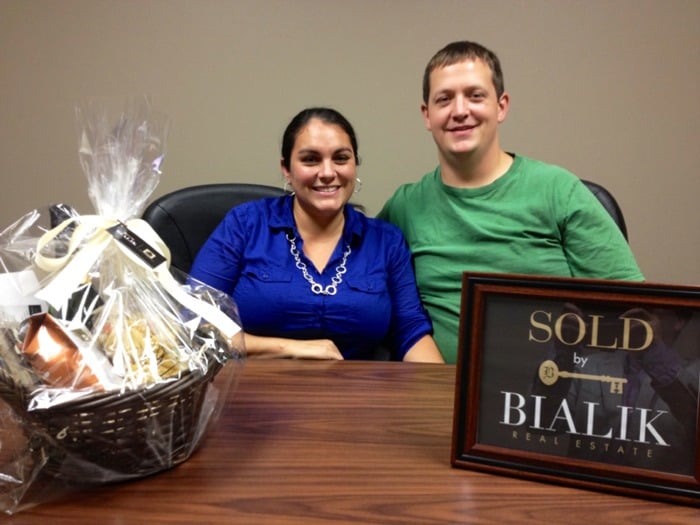 Phil and Trisha smile beside a gift basket and framed sold sign.
