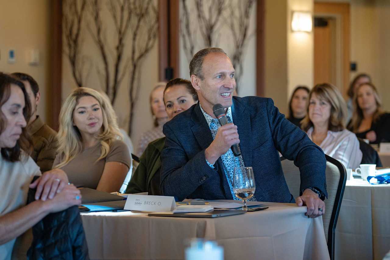 Man speaking into a microphone at a meeting, with attendees seated around him.