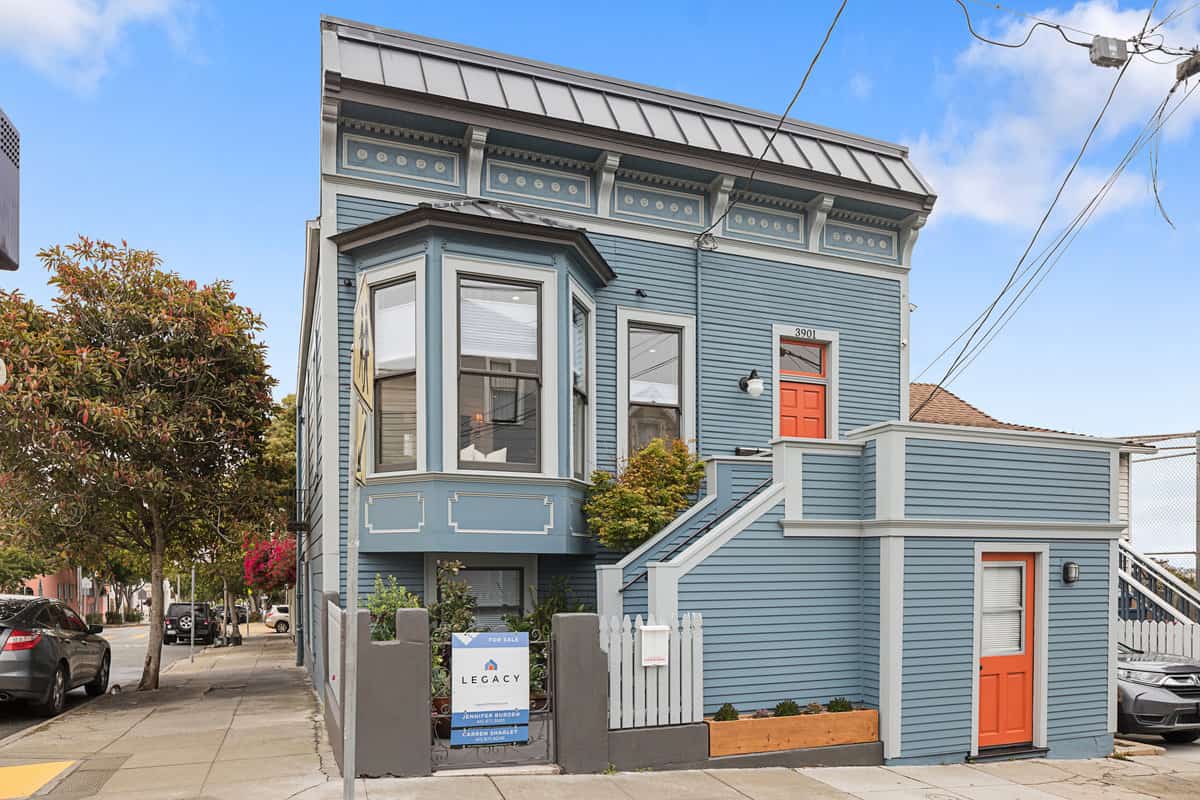 Exterior of a renovated San Francisco Victorian home painted blue, featuring classic bay windows, decorative trim, an orange front door, and a Legacy Real Estate for-sale sign on a sunny city street.