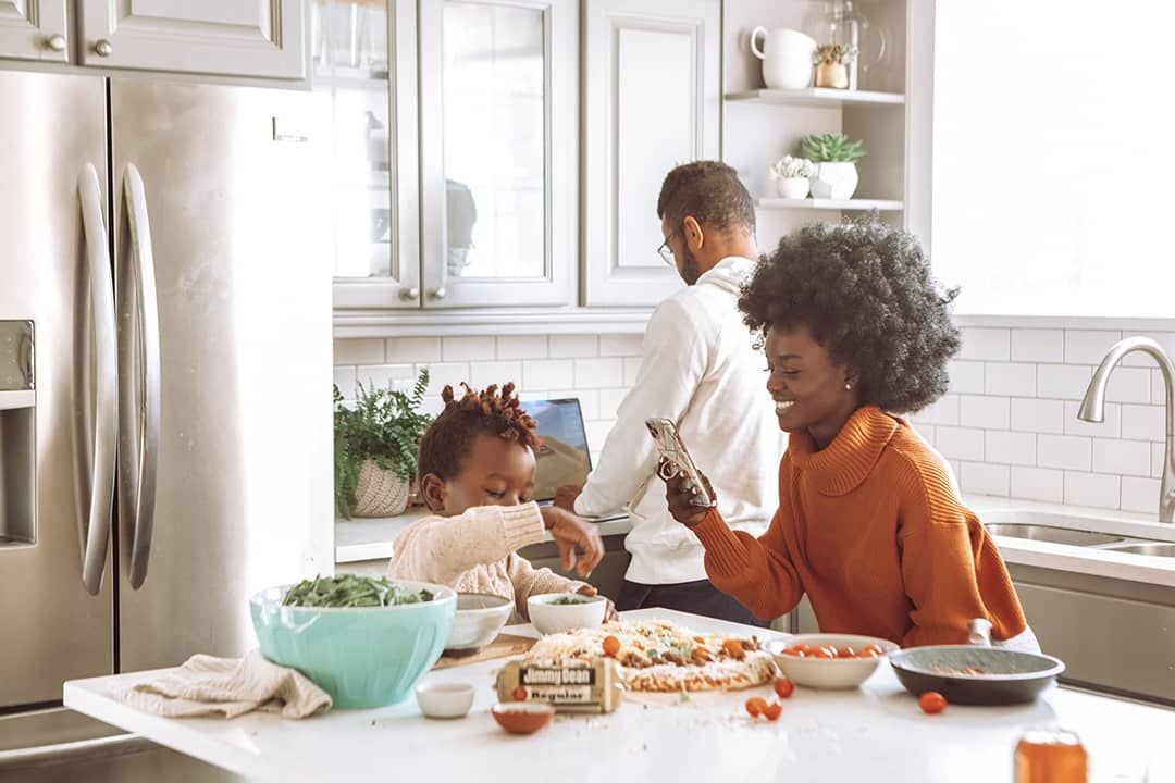 Family prepares food together in a bright kitchen, with a child mixing ingredients at the island.
