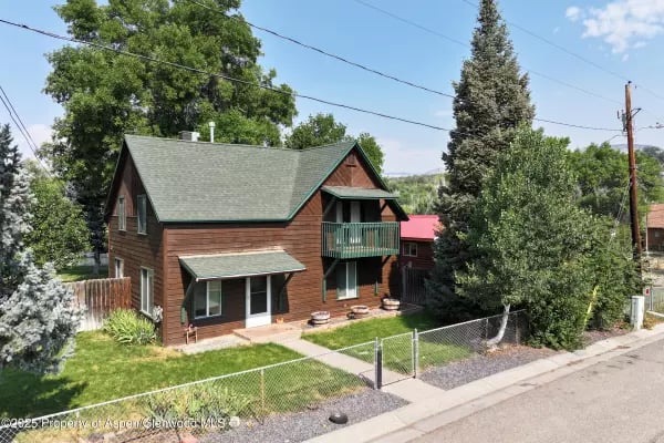 Two-story brown home with a green gabled roof and balcony, behind a fenced lawn and mature trees.