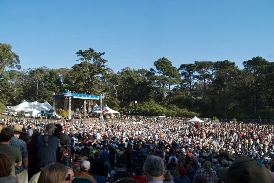 Large crowd seated on grassy hillside facing an outdoor stage with blue signage, surrounded by tall trees under clear blue sky at daytime music festival