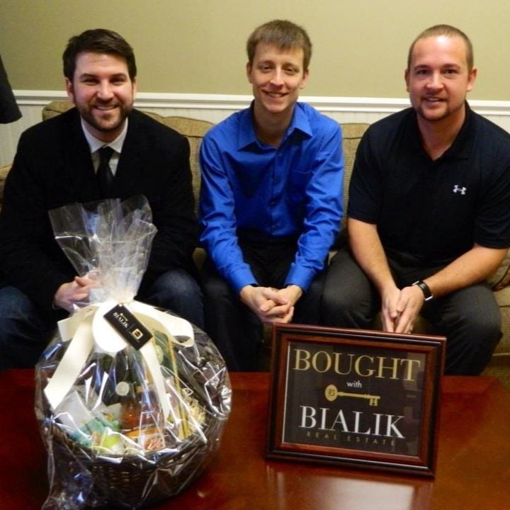 Three men pose with a gift basket and a “Bought with Bialik Real Estate” sign.