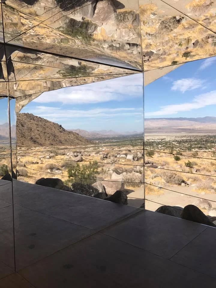Mirrored desert art installation reflecting rocky hills, distant mountains, and blue sky.