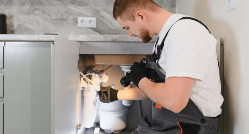 Plumber inspects pipes beneath an open kitchen sink with a flashlight.