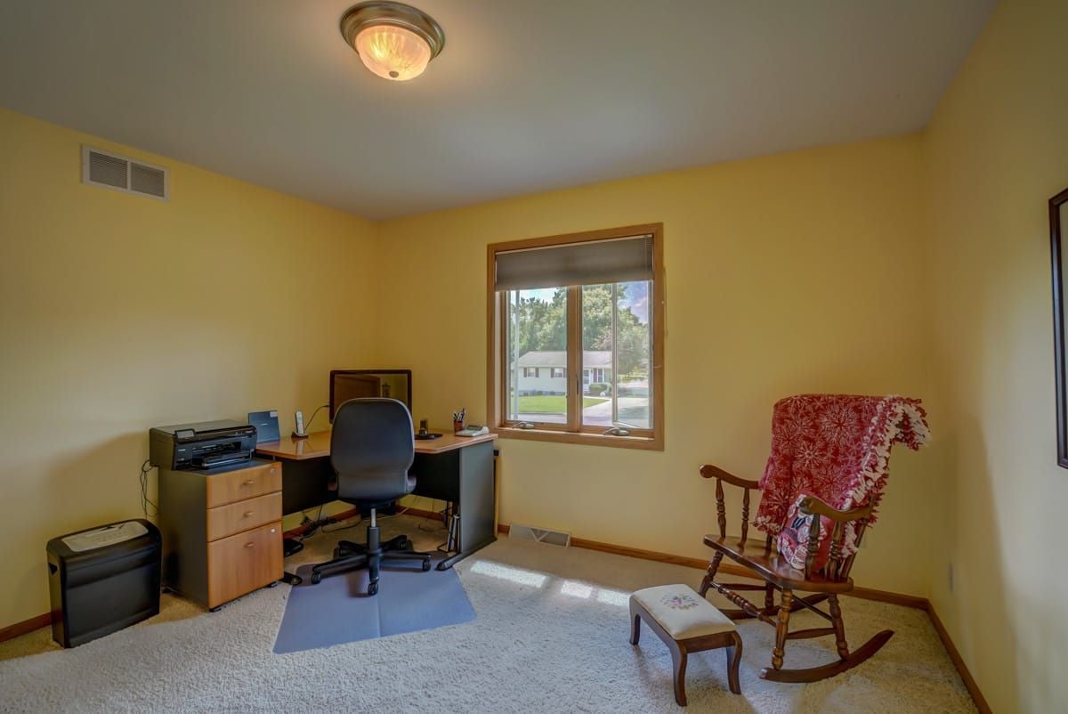 Furnished home office with a desk and computer chair, rocking chair, and sunlit window.