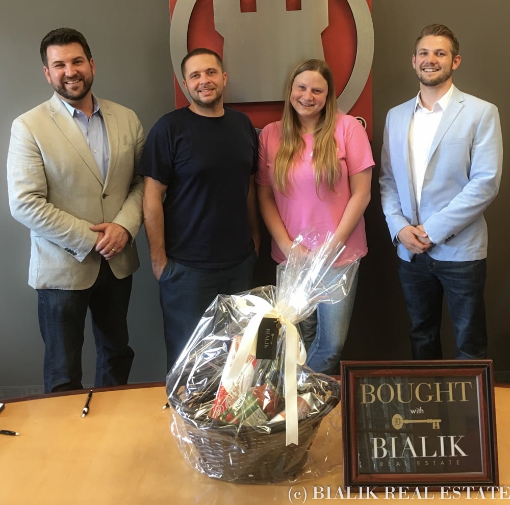 Four adults pose with a gift basket and a Bought with Bialik Real Estate sign.
