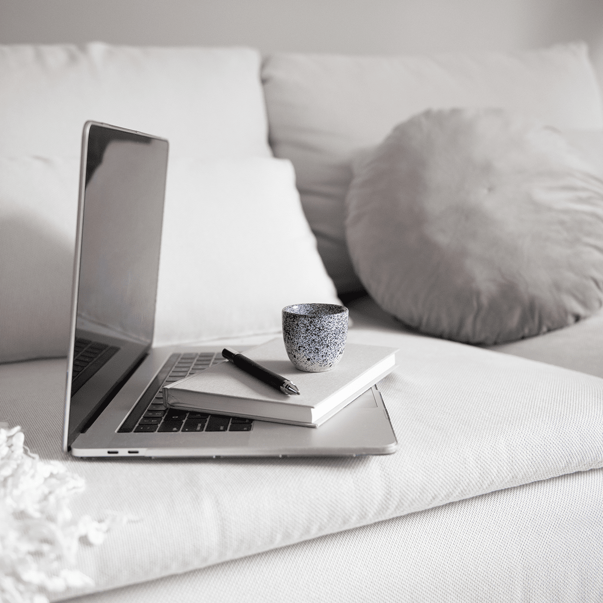 Laptop, notebook, and pen resting on a sofa beside a small cup.