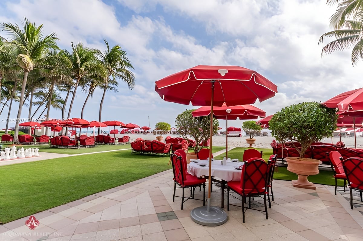 Outdoor dining terrace with red umbrellas and chairs beside a palm-lined beach.