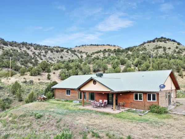 Log-sided home with a pale metal roof and covered patio among shrub-covered hills.