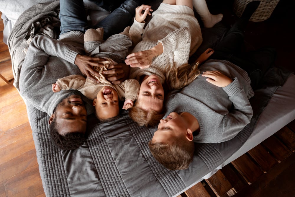 A family of four, including a couple with two children, lying on a cozy bed in warm sweaters, smiling and laughing together, illustrating warmth and togetherness during cold winter weather.
