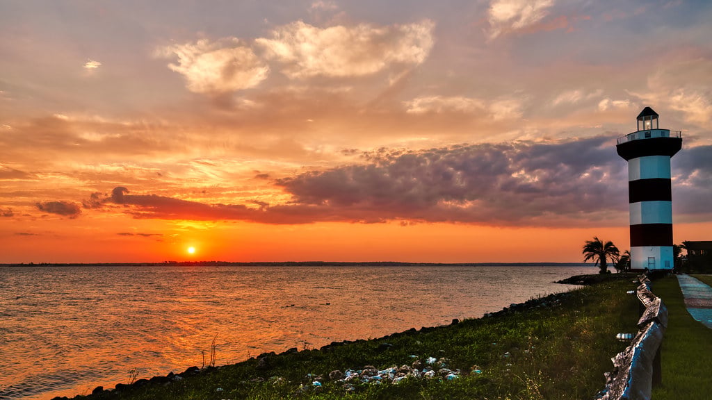 Striped lighthouse beside the water at sunset, with orange clouds and a palm tree.