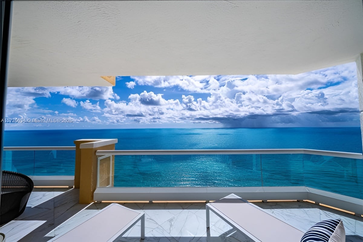Furnished balcony overlooking turquoise ocean, with glass railing and lounge chairs.