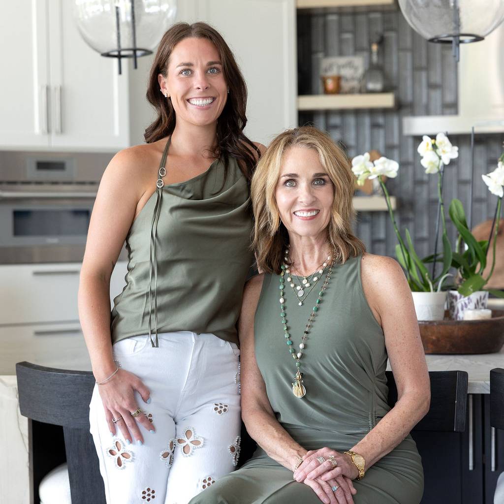 Two smiling women pose in a modern kitchen, one standing and one seated.