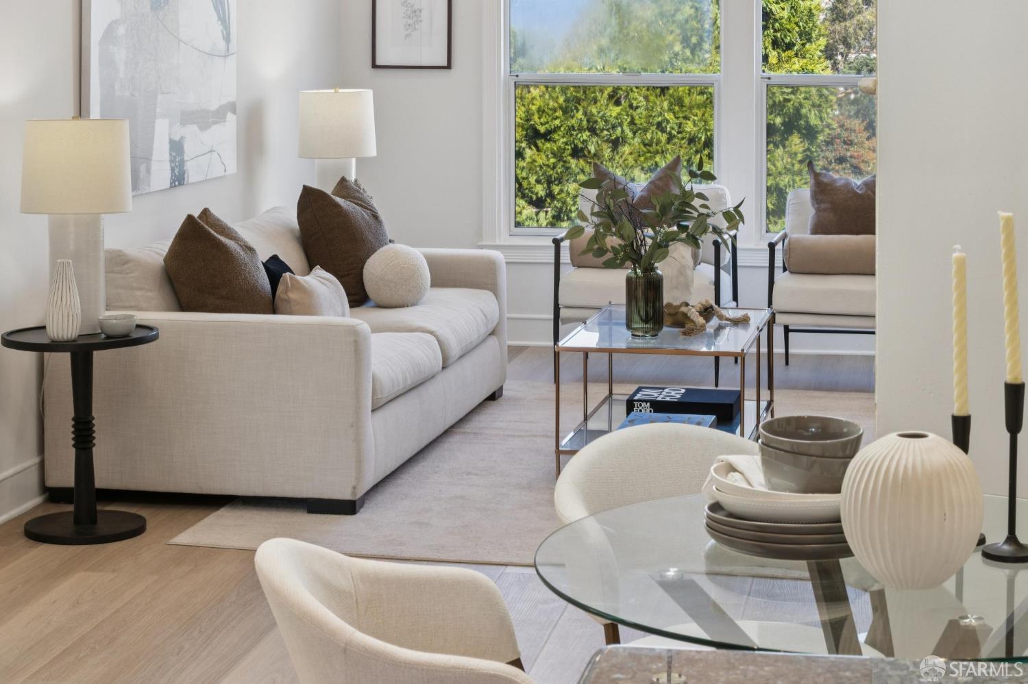 Staged living room with a cream sofa, glass coffee table, and tall windows overlooking greenery.