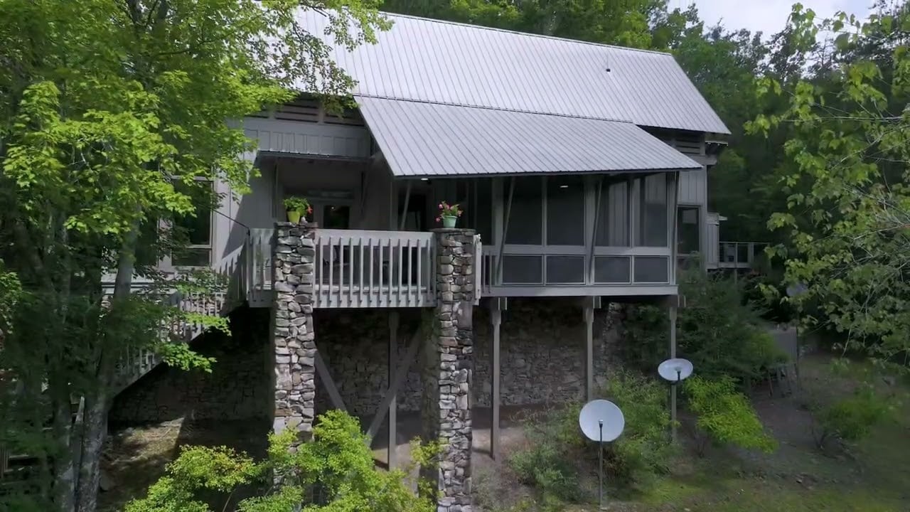 Raised gray house with a metal roof, screened porch, stone pillars, and wooded surroundings.
