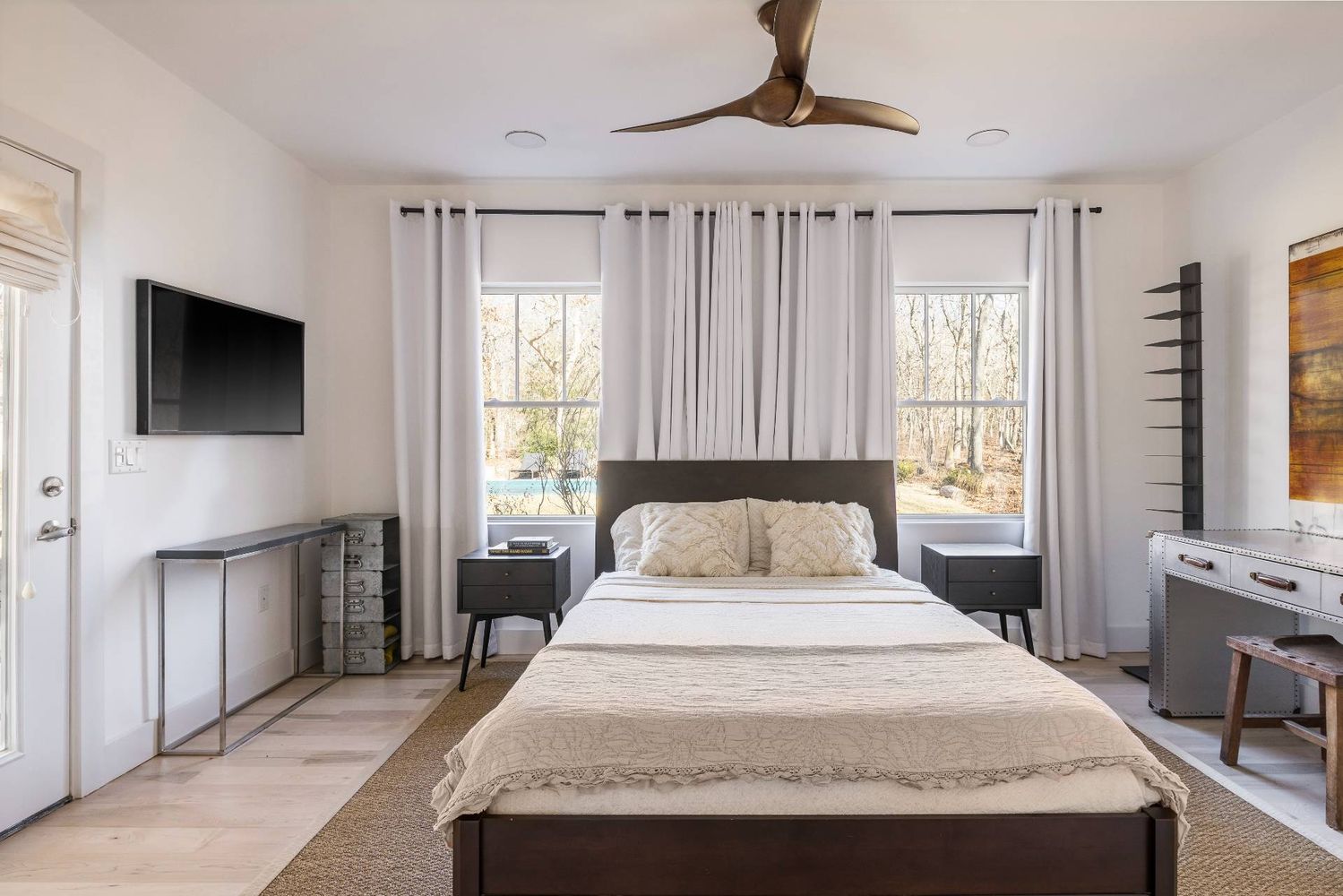 Primary bedroom with a large bed, curtained windows overlooking trees, and a ceiling fan.