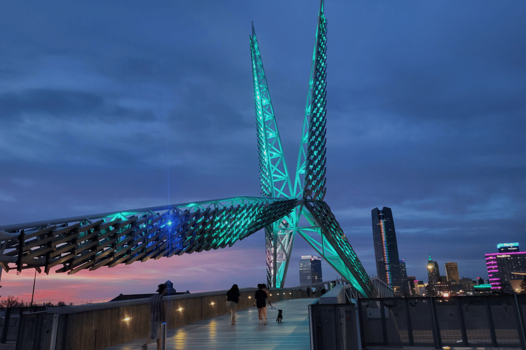 SkyDance Bridge glows turquoise at dusk above pedestrians and the downtown Oklahoma City skyline.