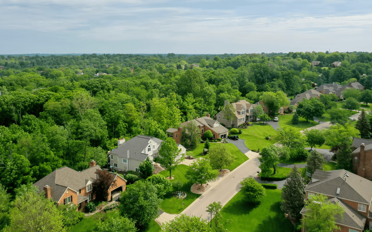 Aerial view of homes along a winding street, surrounded by green lawns and dense woods.