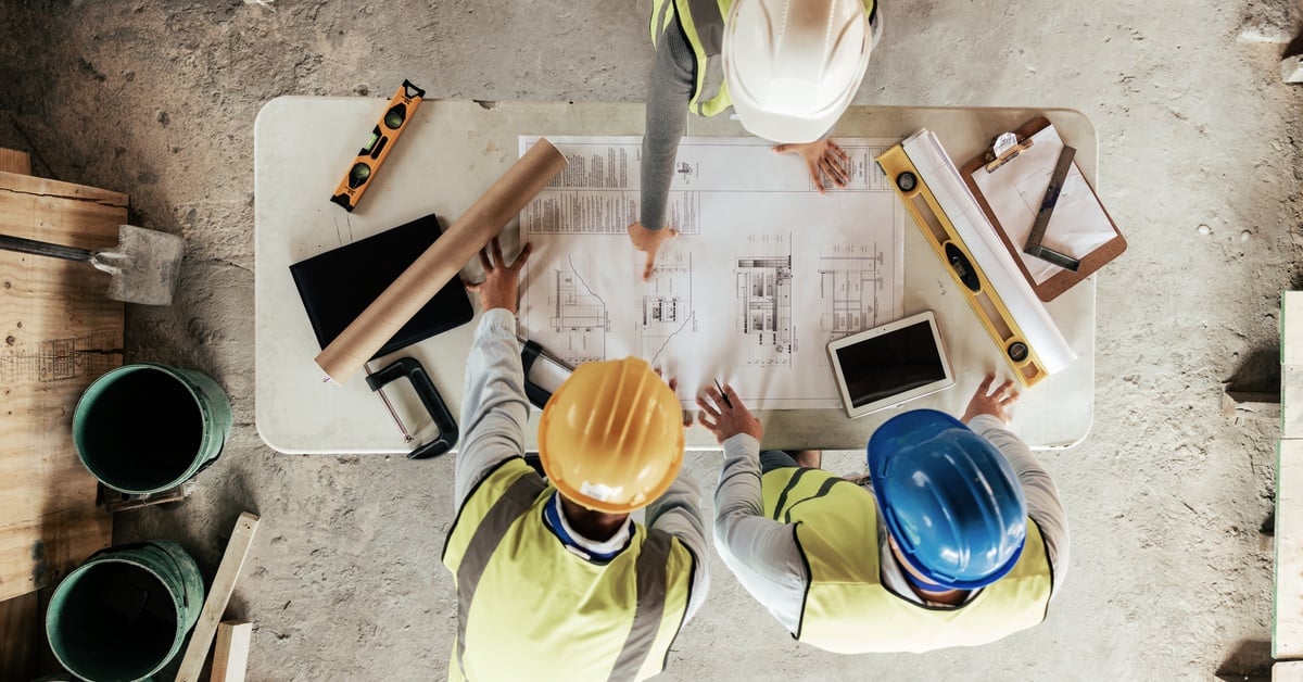 An overhead view of three building contractors in high-visibility vests and hard hats looking at building plans at a site.