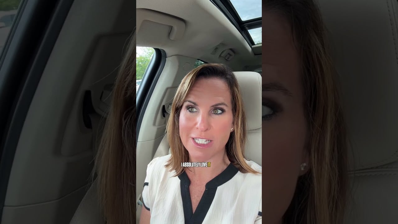 A woman speaking to camera in a car, wearing a white blouse with black trim.
