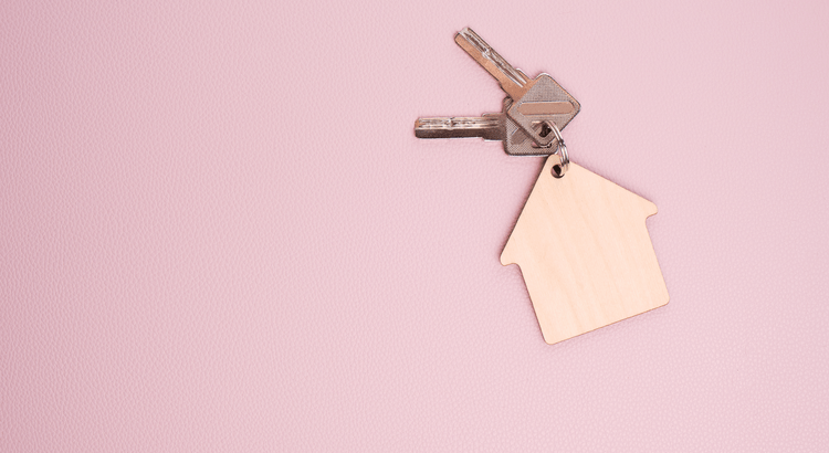 Keys with a house-shaped wooden keychain on a pink background. The simple composition suggests themes of homeownership and real estate.