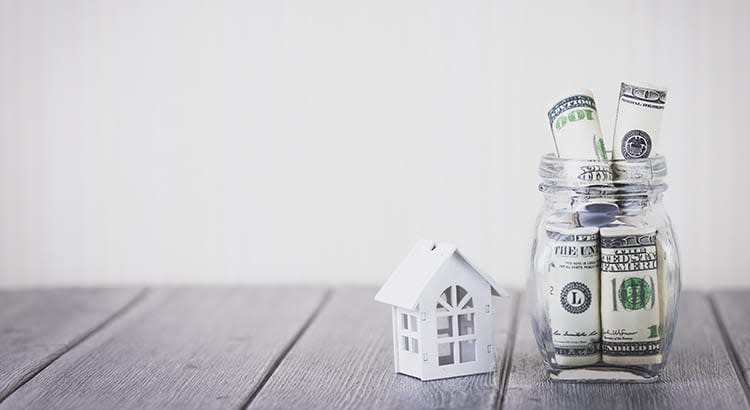 A model house with dollar bills in a jar which emphasizes savings, real estate investment, or financial planning, with a small house model next to rolled-up cash in a glass jar.