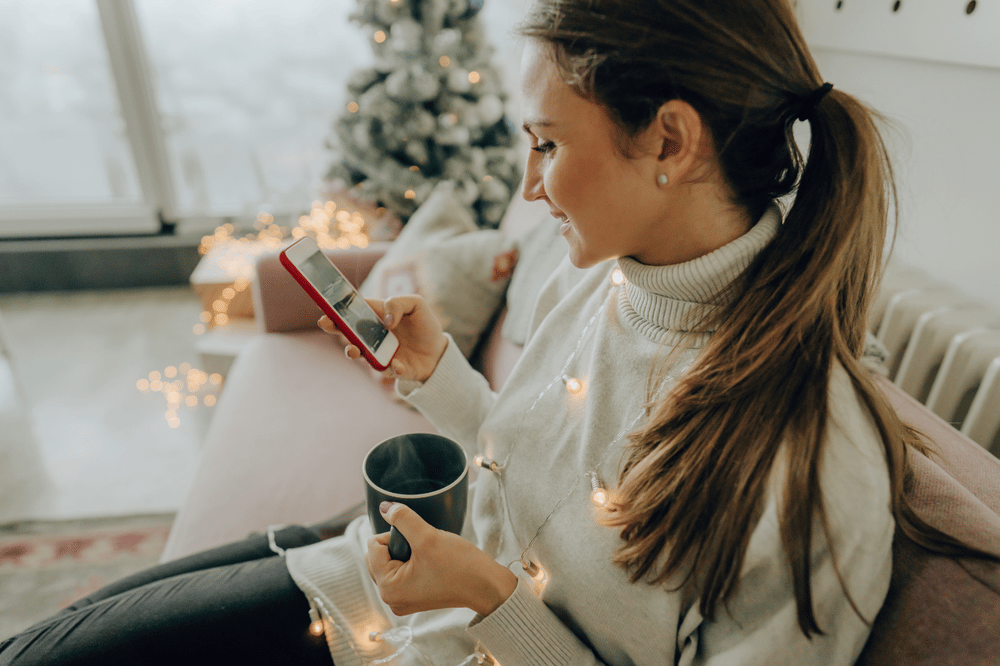 Woman checks her phone and holds a mug beside a lit Christmas tree.