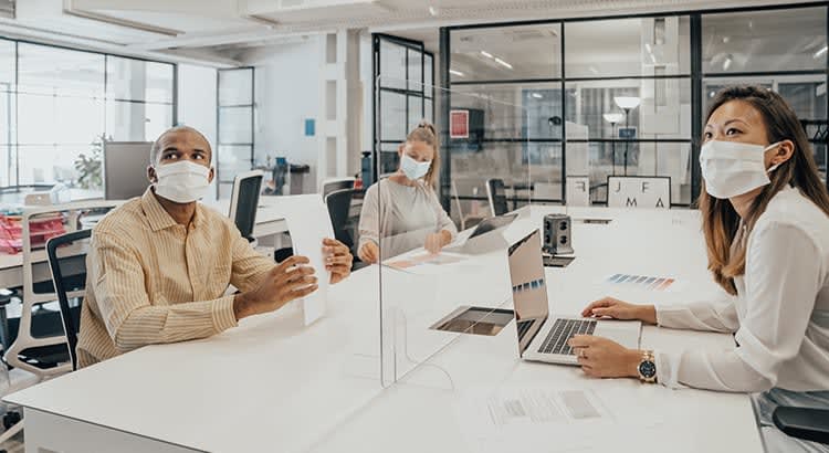 a modern office setting where three individuals are seated at a large white table, each engaged in work-related activities.