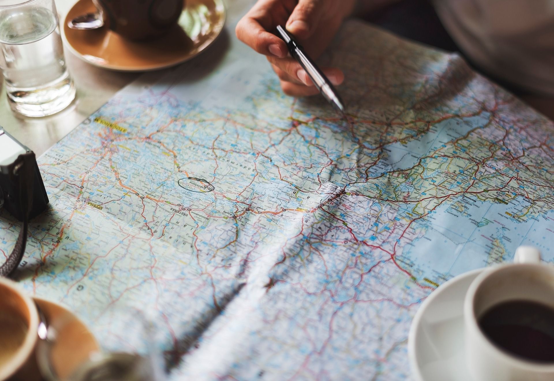 A hand points to a road map with a pen beside coffee cups.