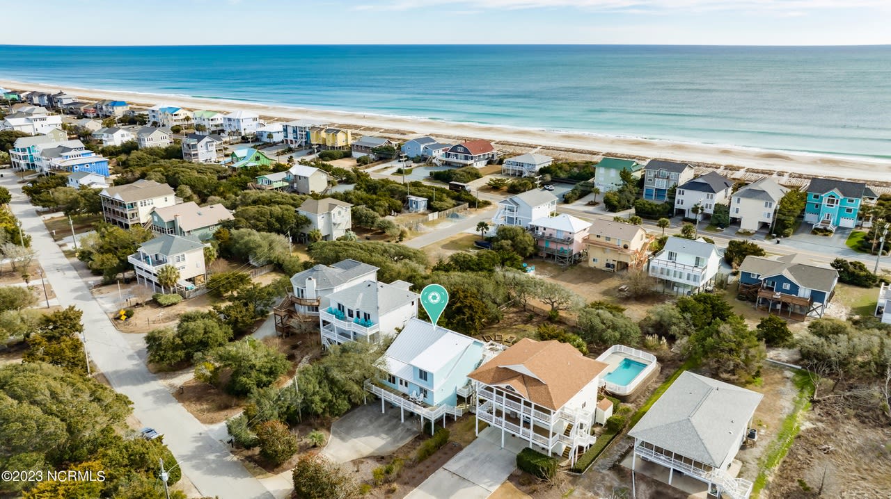 Topsail Beach homes for sale. A row of colorful houses with private pools, balconies, patios, trees, and shrubs,  on a sandy beach next to a turquoise ocean.