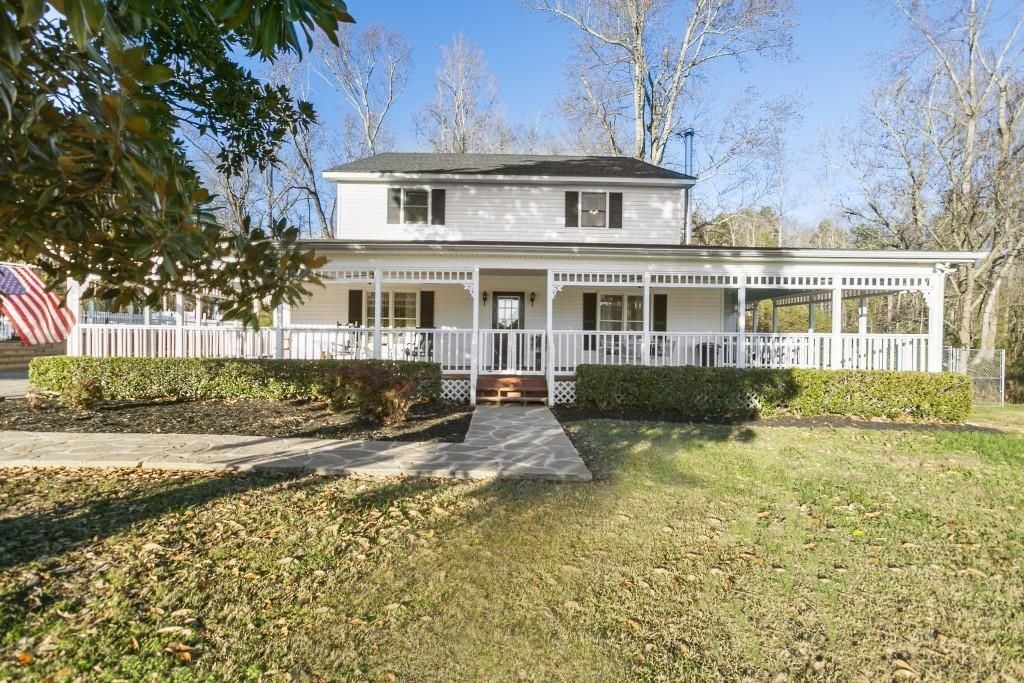 White two-story home with a wraparound porch, stone walkway, and American flag.