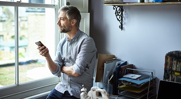 A man with a beard sits near a window, possibly reflecting while looking outside, with a relaxed posture, suggesting a quiet, contemplative moment.