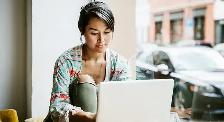 A woman sitting and using a laptop, with a thoughtful expression. She appears to be in a casual setting, possibly working or studying.