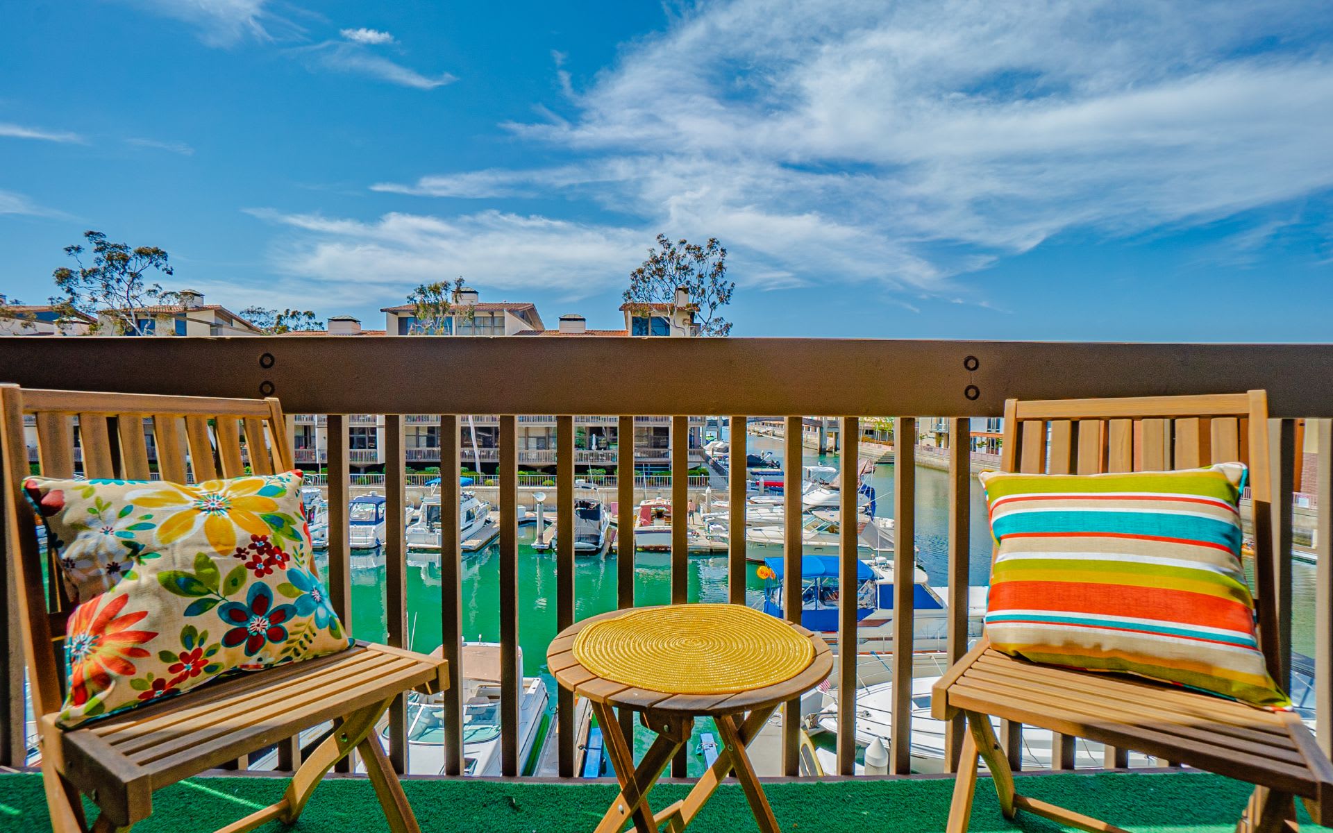 Furnished balcony overlooking a marina, with colorful cushions, wooden chairs, and boats on turquoise water.