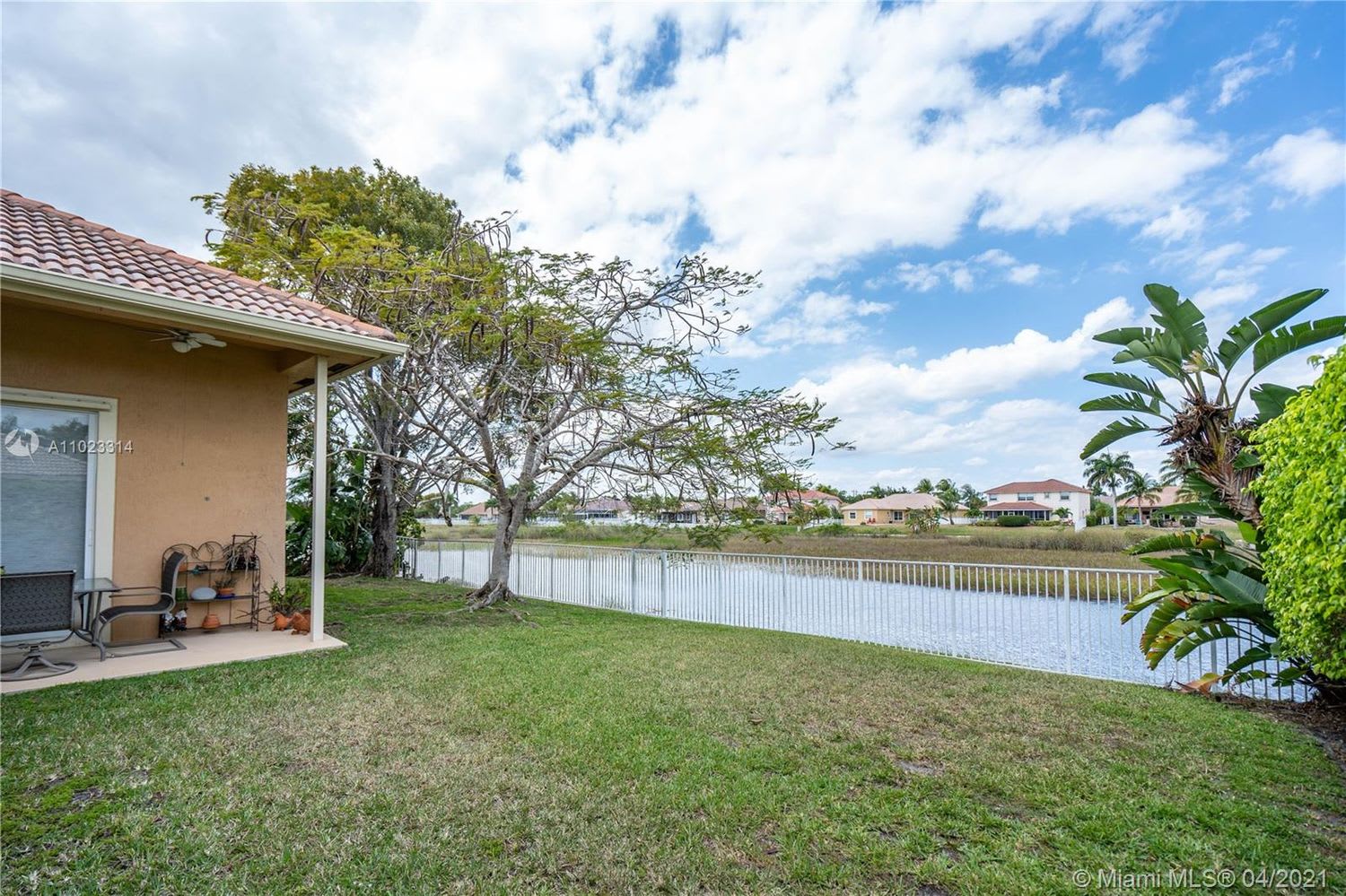 Grassy backyard beside a canal, with a covered patio, white fence, and neighboring homes across the water.