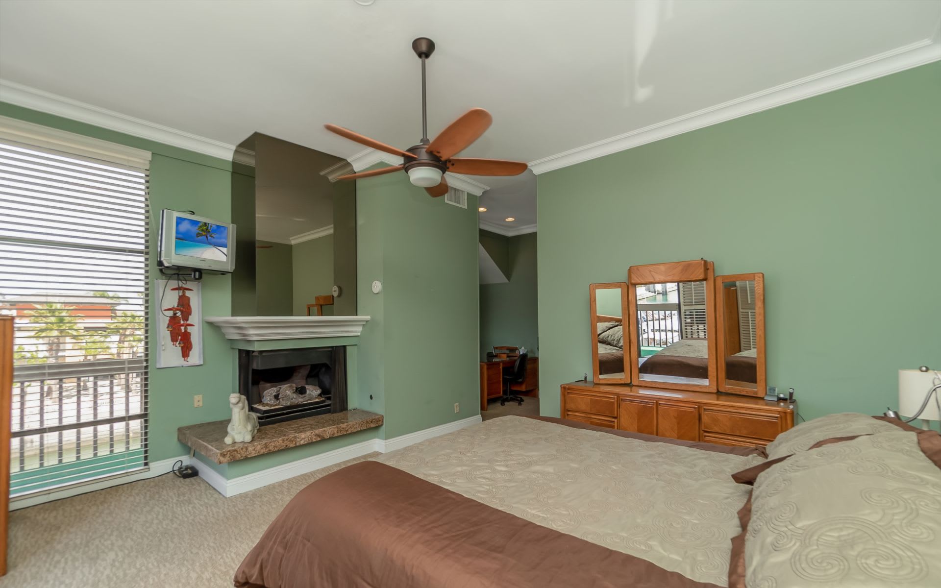 Bedroom with sage green walls, a fireplace, ceiling fan, and large window with blinds.