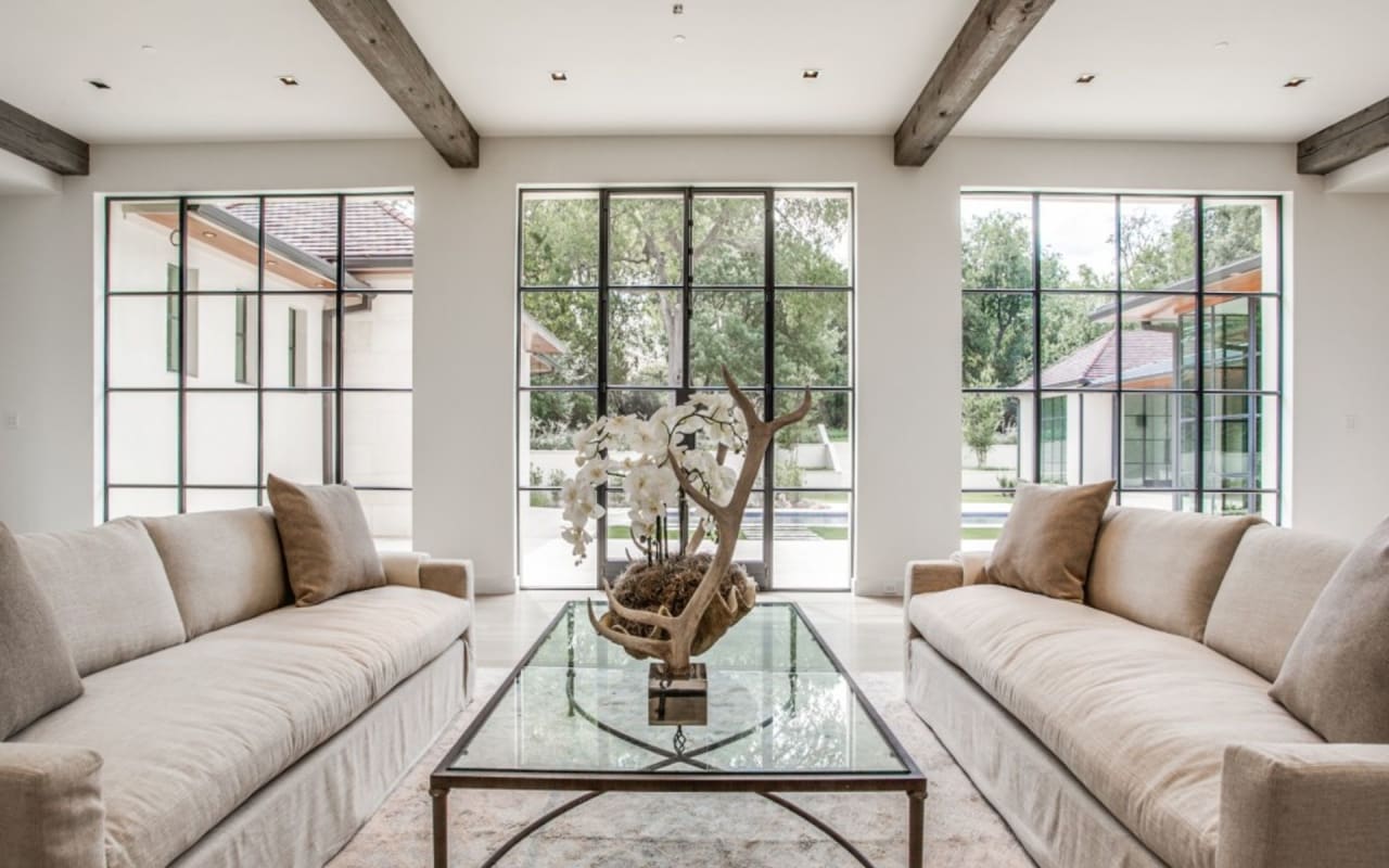 Inviting seating area with white sofas, overlooking the patio and backyard through beautiful oversized black-framed windows.