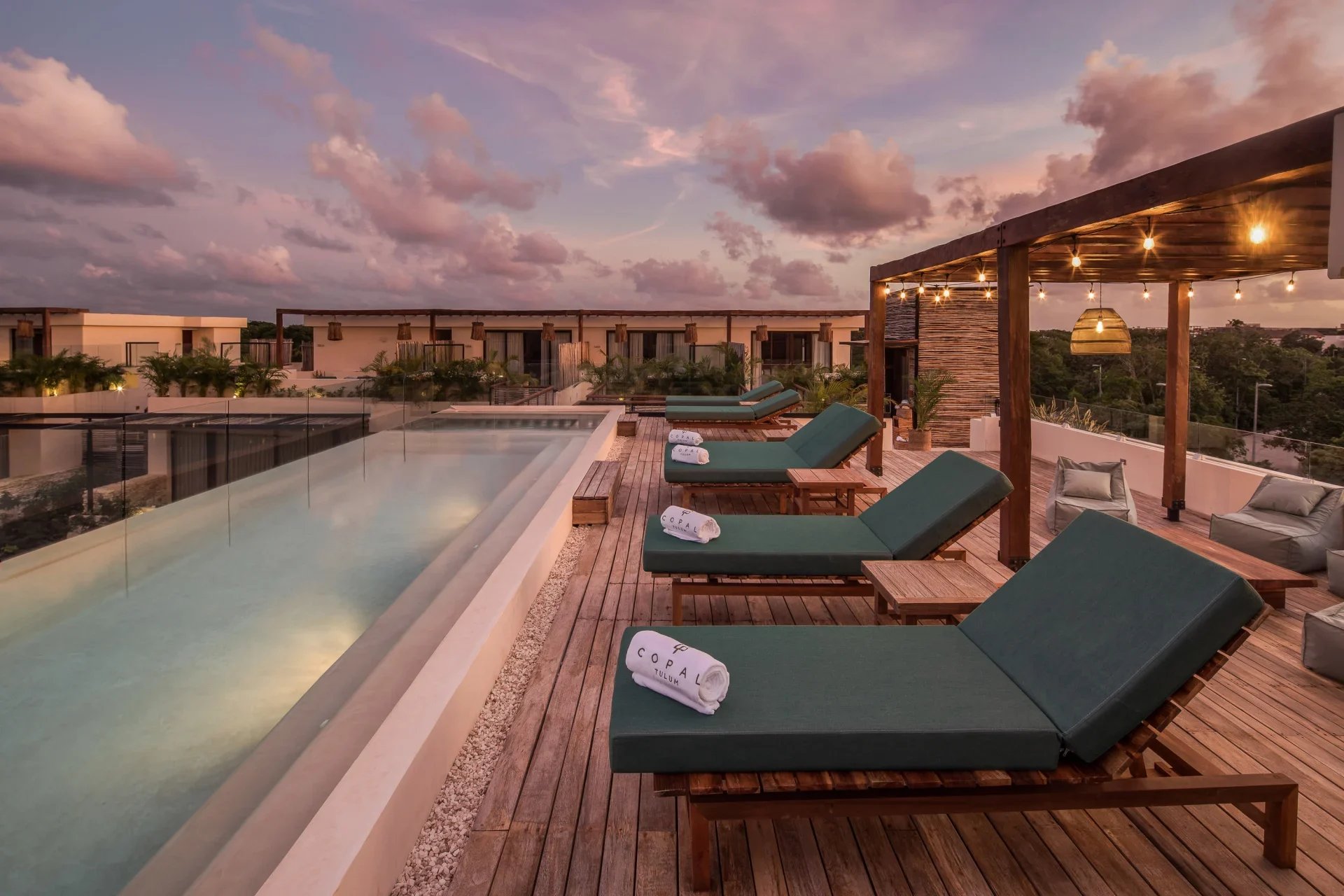 Rooftop pool at dusk with green loungers and a wooden pergola strung with lights.