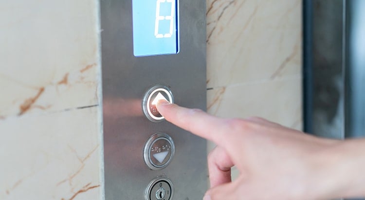 A close-up image of a finger pressing an elevator button in a metallic and marble interior. The button is lit up, indicating it has been pressed.