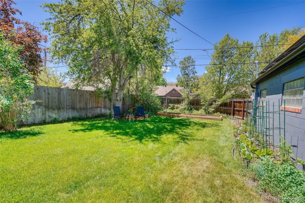 Backyard with a broad lawn, mature trees, wooden fencing, patio chairs, and a raised garden bed.