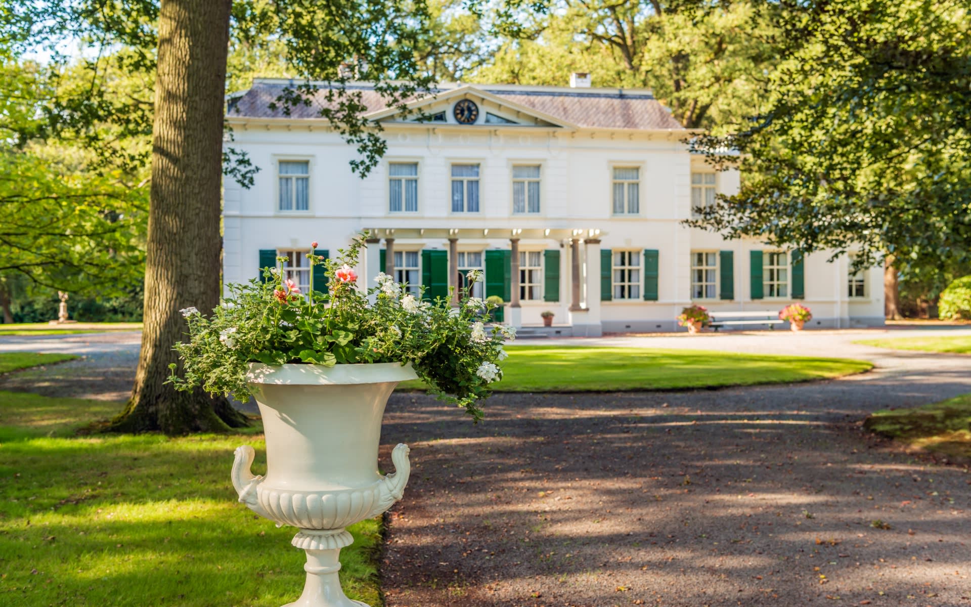  A grand, two-story white mansion with dark green shutters stands prominently in the background, partially obscured by lush green trees. 