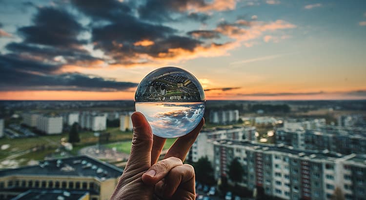 A hand holding a transparent glass orb (likely a crystal ball), which reflects an inverted view of an urban landscape beneath a dramatic, cloud-filled sky during sunset. The buildings and horizon visible through the crystal ball are part of a cityscape, with multiple apartment buildings and residential blocks.