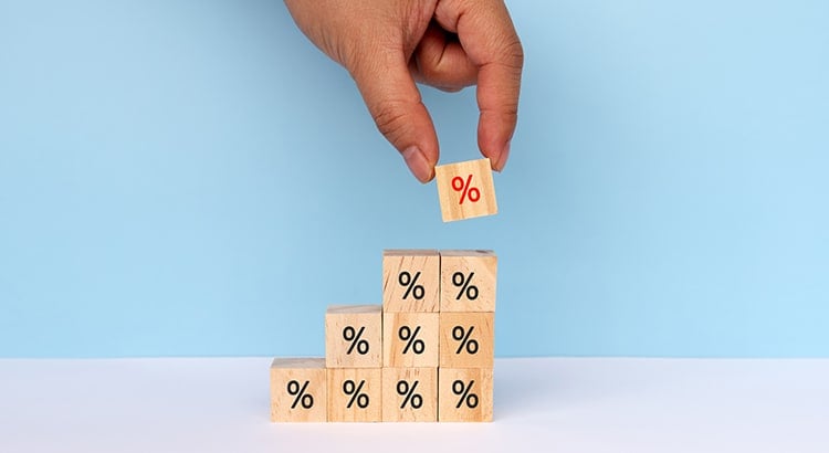 A hand placing a wooden block with a percentage symbol on top of a stack of similar blocks. The blocks are arranged to form a staircase, suggesting growth or increase.