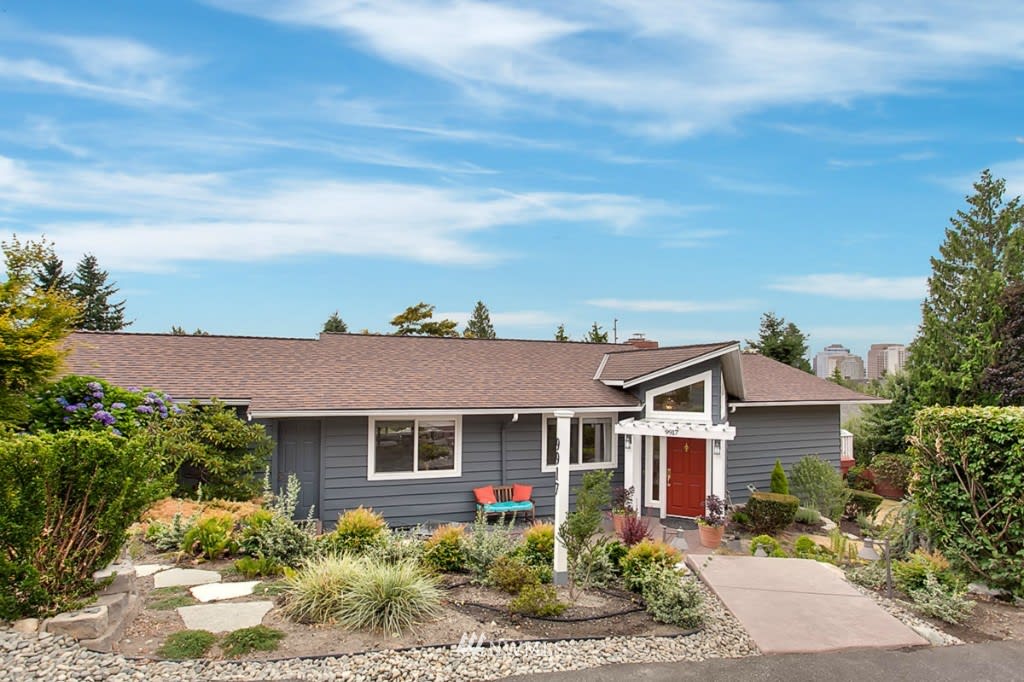 Gray single-story house with a red front door, landscaped yard, and distant city skyline.