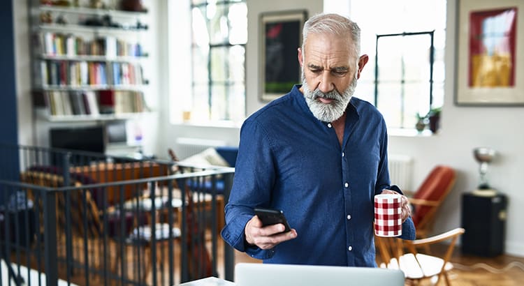 An older man looking at a his phone in one hand and a mug in the other hand.