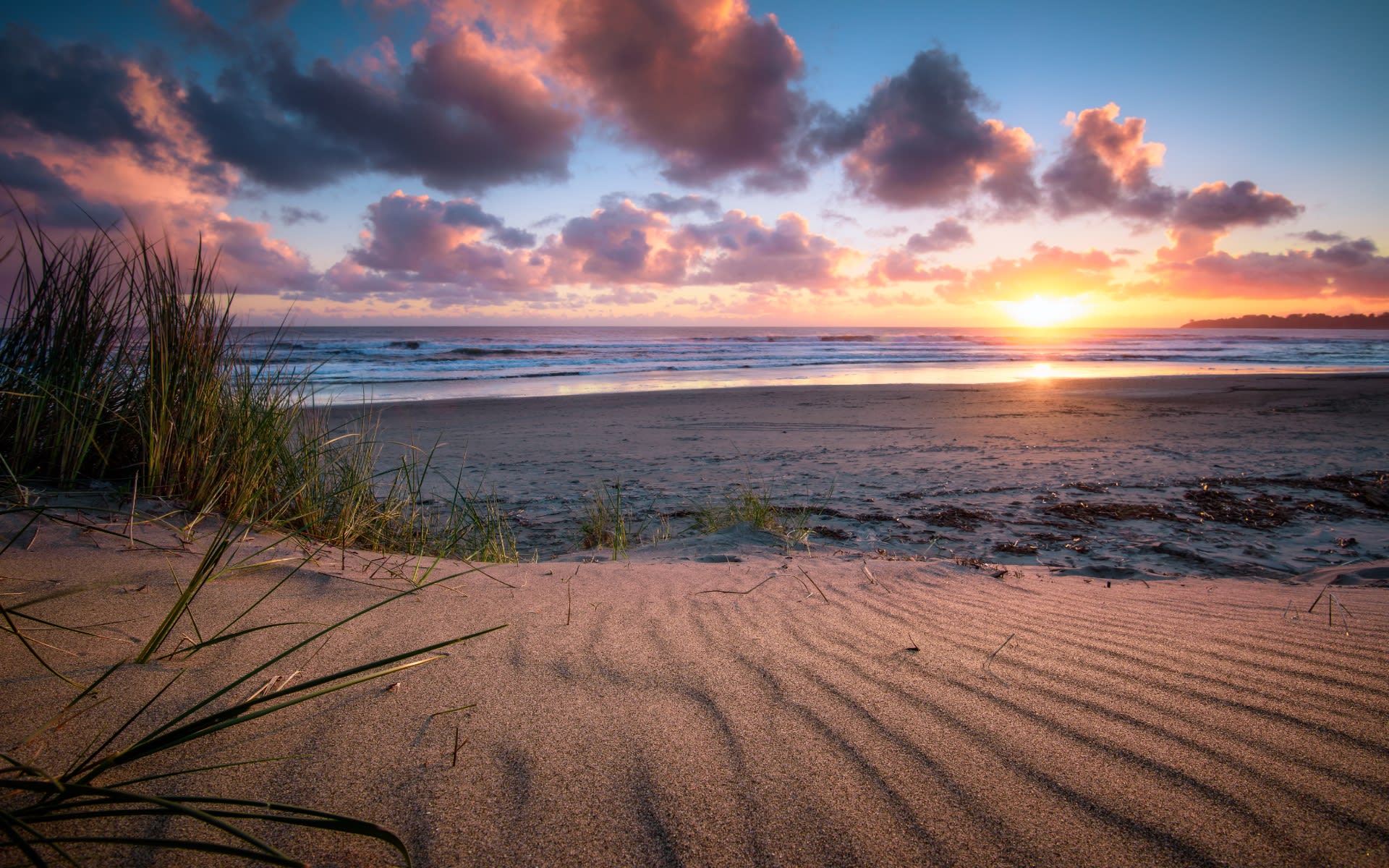 Sunset over a sandy beach with dune grass, ocean waves, and colorful clouds in the sky.