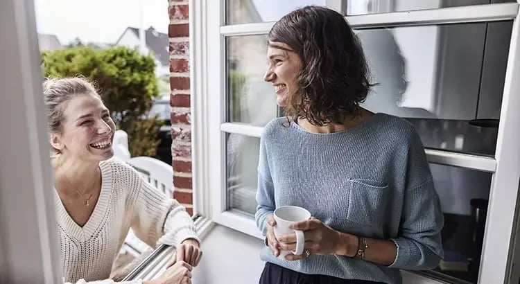 The woman on the left is leaning on a window sill and is laughing. The woman on the right is holding a white coffee mug and is smiling warmly. 